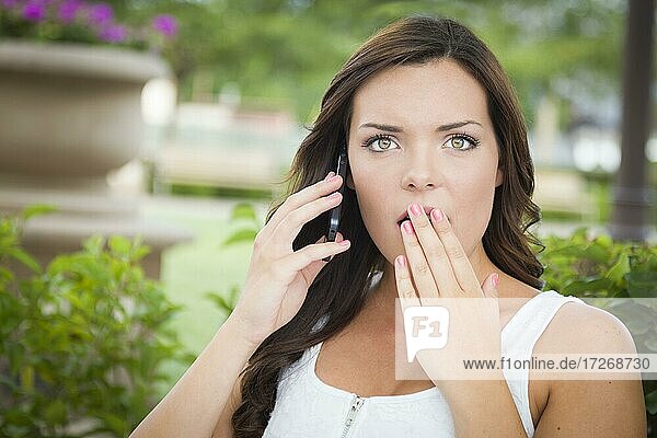 Shocked young adult female talking on cell phone outdoors on bench