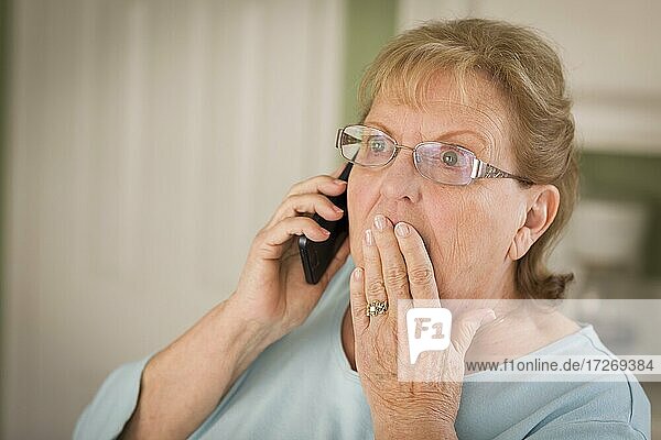 Shocked senior adult woman on cell phone with hand over mouth in kitchen
