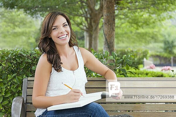 Attractive young adult female student on bench outdoors with books and pencil