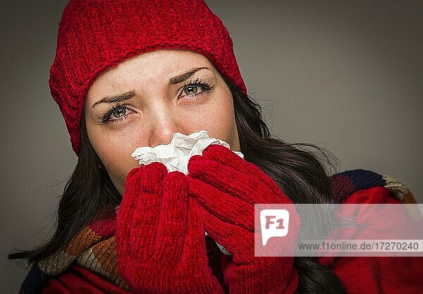 Kranke gemischtrassige Frau mit Wintermütze und Handschuhen  die sich die wunde Nase mit einem Taschentuch putzt