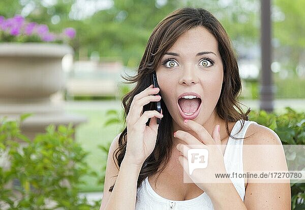 Schockierte junge adultes Weibchen  die im Freien auf einer Bank mit einem Handy telefoniert