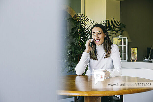 Junge Frau  die mit ihrem Handy telefoniert und dabei Kaffee trinkt  während sie zu Hause sitzt