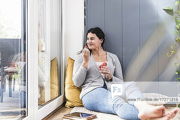 Smiling young woman eating strawberry while sitting by window at home