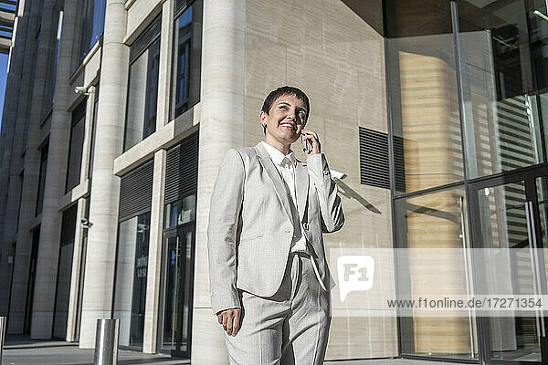 Lächelnde Geschäftsfrau  die mit einem Mobiltelefon spricht  während sie vor einem modernen Bürogebäude steht