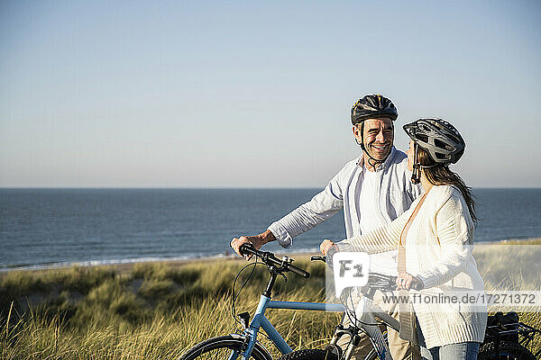 Happy couple in cycling helmets standing with bicycle at beach against clear sky