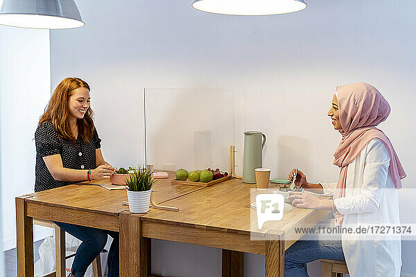 Lächelnde Frauen beim Essen  die an einem Tisch mit Bildschirmteilung im Büro sitzen