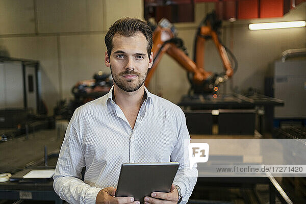 Confident young man with digital tablet standing against robotic arm at factory