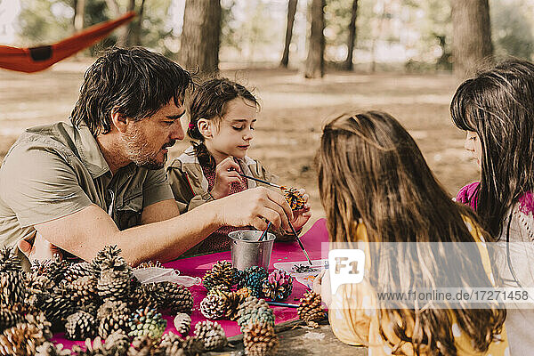 Mature man with daughters decorating while coloring pine cones at picnic table in park