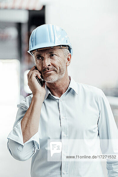 Männlicher Ingenieur mit Schutzhelm schaut weg  während er mit einem Smartphone in der Industrie spricht