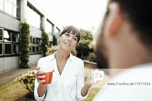 Geschäftsfrau mit Pony  die eine Kaffeetasse in der Hand hält  während sie einem männlichen Kollegen während einer Diskussion in der Pause zuprostet