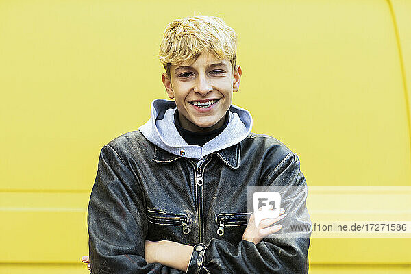 Smiling blond teenage boy standing with arms crossed against yellow van in city