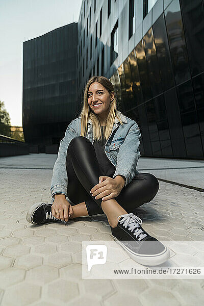 Lächelnde junge Frau  die wegschaut  während sie auf dem Fußweg vor einem modernen Gebäude sitzt