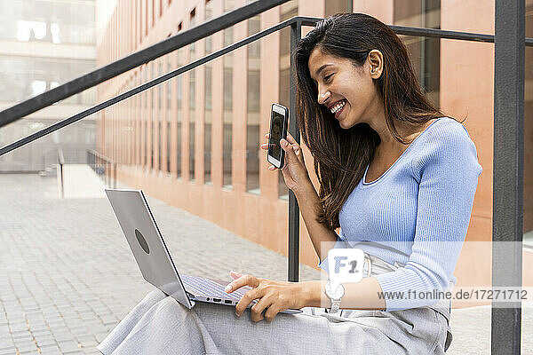 Lächelnde Geschäftsfrau zeigt ihr Smartphone bei einem Videotelefonat über einen Laptop während der COVID-19