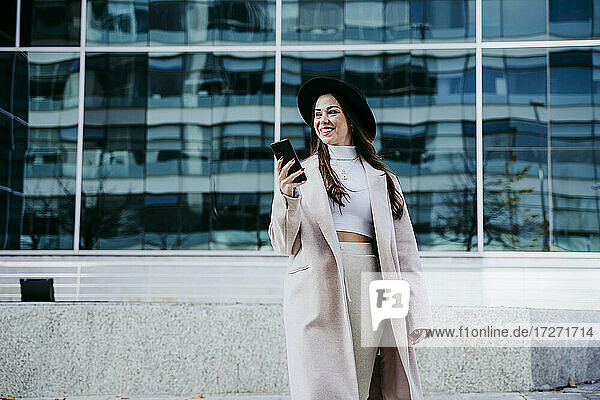 Lächelnde Frau mit Smartphone schaut weg  während sie an einem Gebäude steht