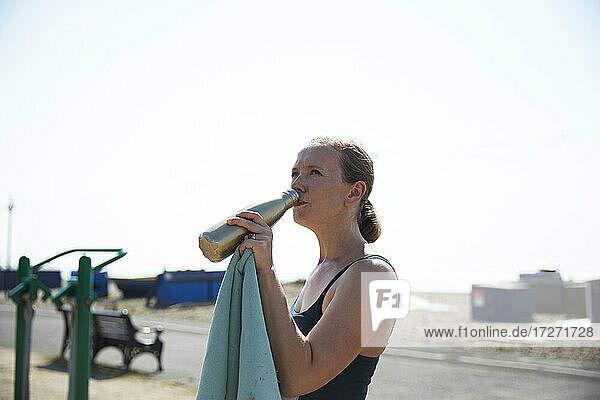 Reife Frau trinkt Wasser  während sie in einem Fitnessstudio im Freien an einem sonnigen Tag steht