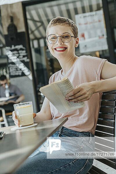Lächelnde erwachsene Frau  die ein Buch liest  während sie auf dem Bürgersteig sitzt und Kaffee trinkt  an einem sonnigen Tag