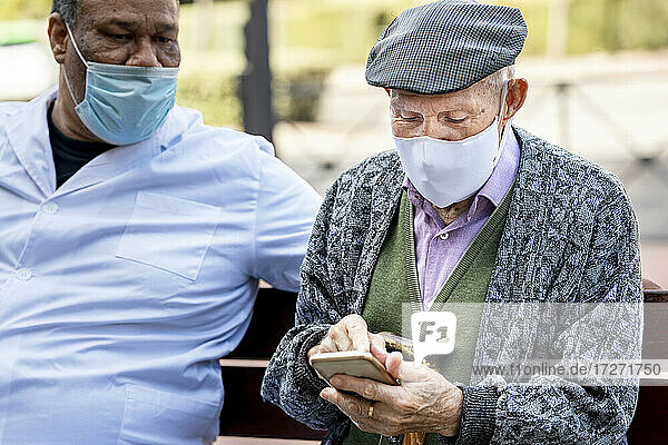 Älterer Mann  der ein Smartphone benutzt und einen Gesichtsschutz trägt  sitzt mit einem älteren Mann auf einer Bank während COVID-19