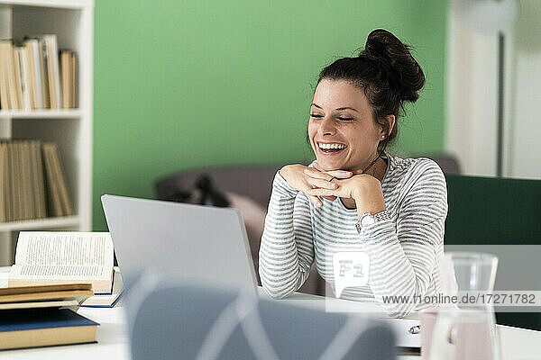 Frau lachend mit Hand am Kinn bei der Benutzung eines Laptops  während sie zu Hause sitzt