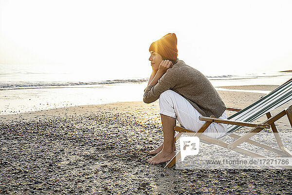 Nachdenkliche junge Frau sitzt auf einem Klappstuhl und blickt bei Sonnenuntergang auf den Strand und den klaren Himmel