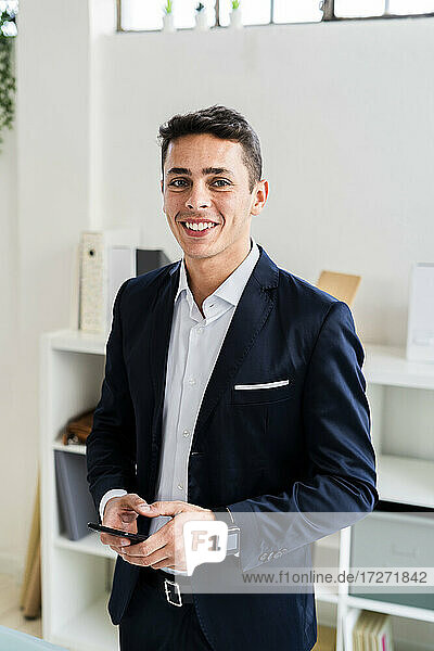 Lächelnder  gutaussehender Geschäftsmann  der ein Smartphone hält  während er an einem kreativen Arbeitsplatz steht