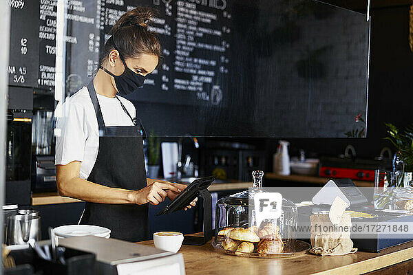 Weiblicher Barista mit Gesichtsmaske bei der Verwendung eines digitalen Tablets an der Küchentheke eines Cafés