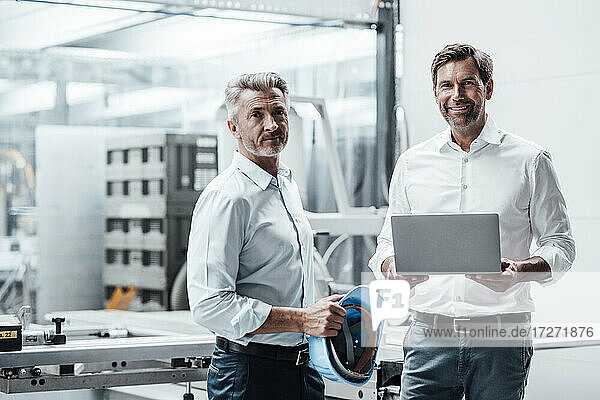 Lächelnder männlicher Ingenieur mit Laptop  der neben einem Kollegen mit Schutzhelm in einer Fabrik steht