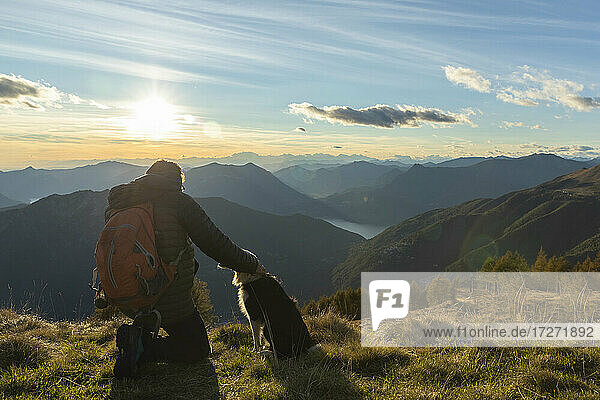 Hiker kneeling while stroking dog at mountain peak during sunset