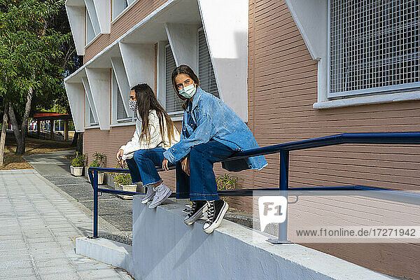 Female friends looking away wearing protective face mask while sitting on railing during sunny day