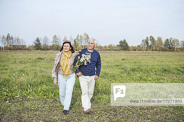Senior couple with flowers walking on field against sky