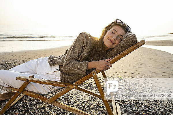 Lächelnde schöne junge Frau  die sich auf einem Klappstuhl ausruht  während sie am Strand bei Sonnenuntergang entspannt