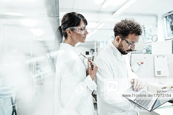 Eine Wissenschaftlerin steht neben einem männlichen Kollegen  der einen Laptop im Labor benutzt