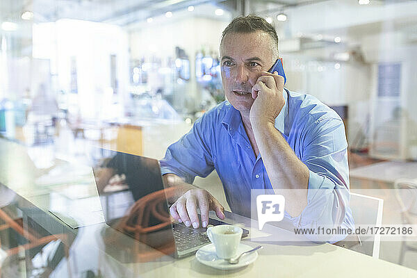 Männlicher Berufstätiger  der mit Laptop und Kaffee in einem Café sitzt und mit seinem Handy telefoniert