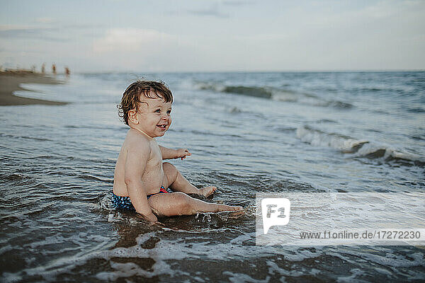 Männliches Kleinkind genießt das Sitzen im Wasser am Strand bei Sonnenuntergang
