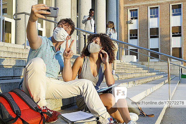 Männlicher Freund gestikuliert Frieden  während er ein Selfie mit einem Universitätsstudenten mit Schutzmaske auf einer Treppe in der Stadt macht