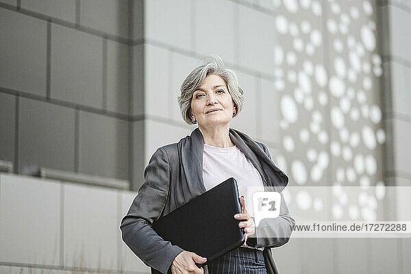 Mature businesswoman holding laptop while standing with hand in pocket against wall