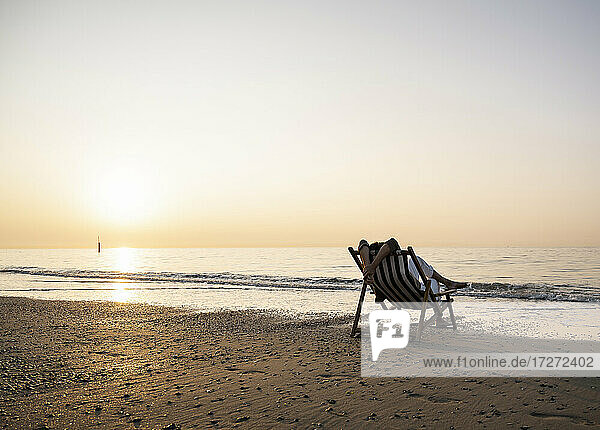 Frau  die sich auf einem Klappstuhl am Strand vor einem klaren Himmel bei Sonnenuntergang entspannt