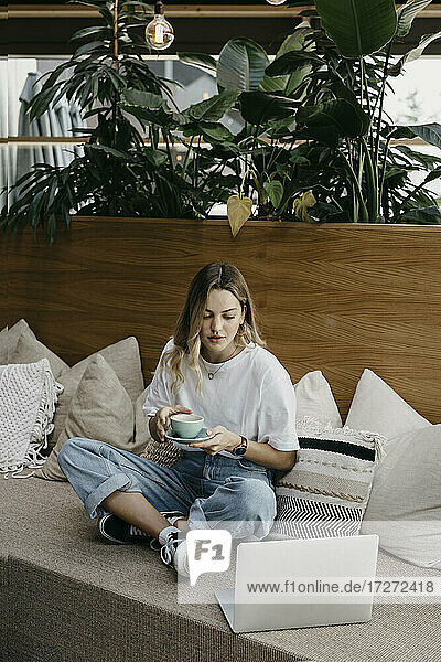 Junge Frau hält eine Kaffeetasse und benutzt einen Laptop auf dem Sofa