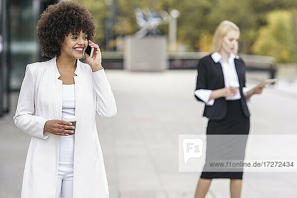 Businesswoman talking on mobile phone while standing with colleague in background on footpath