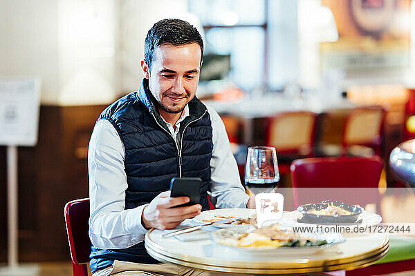 Smiling young man text messaging on smart phone while sitting in restaurant