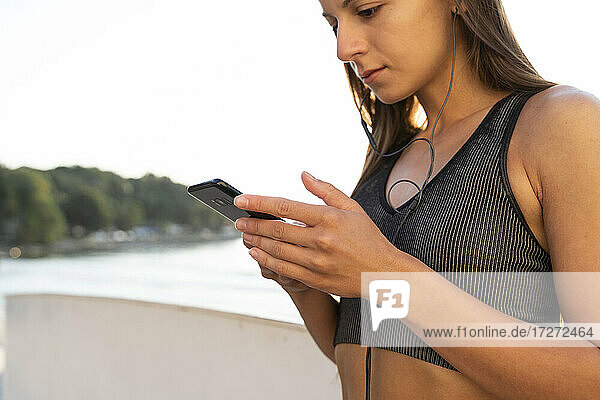 Fitness-Frau hört Musik über Kopfhörer mit Smartphone  während sie bei Sonnenaufgang am Meer steht