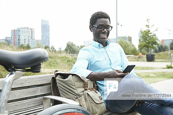Lächelnder Mann  der ein Smartphone benutzt und über Kopfhörer Musik hört  sitzt auf einer Bank in der Stadt
