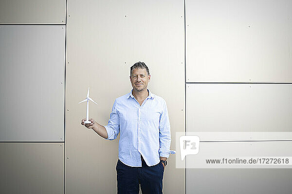 Geschäftsmann lächelnd mit Händen in den Taschen  der eine Windkraftanlage an der Wand hält