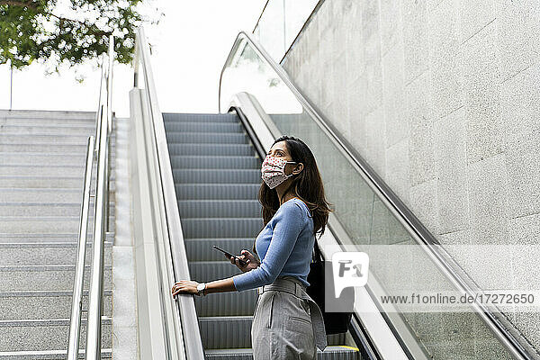 Nachdenkliche Frau  die mit ihrem Handy nach oben schaut  während sie auf der Rolltreppe steht  während COVID-19