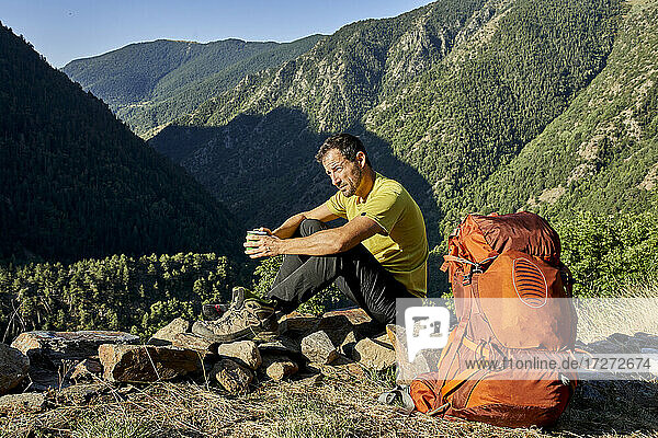 Man looking away while sitting on stone holding coffee cup in forest during sunny day