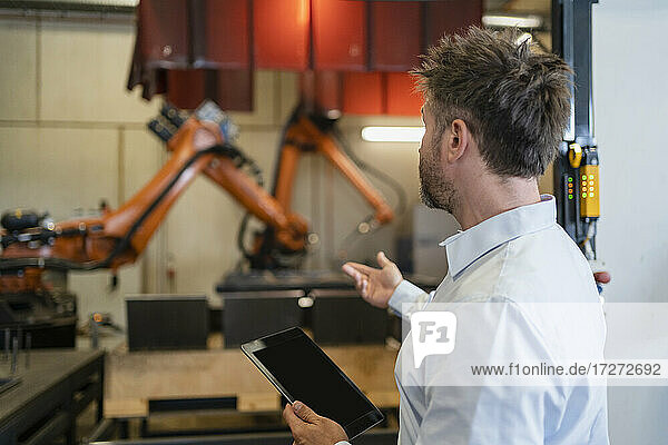 Mature businessman with digital tablet gesturing toward robotic arm machine while standing at factory