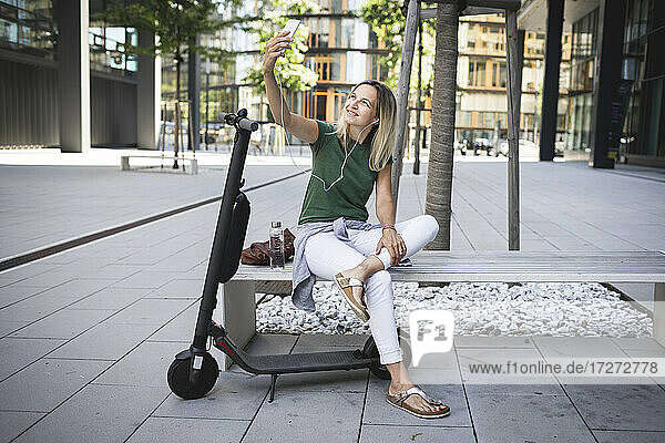 Lächelnd Mitte erwachsene Frau hören Musik und nehmen selfie beim Sitzen auf Bank in der Stadt