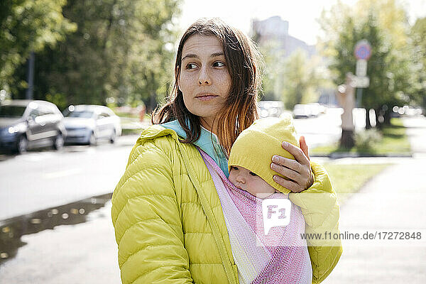 Mutter hält ihr in eine Decke eingewickeltes Baby  während sie an einem sonnigen Tag auf der Straße steht