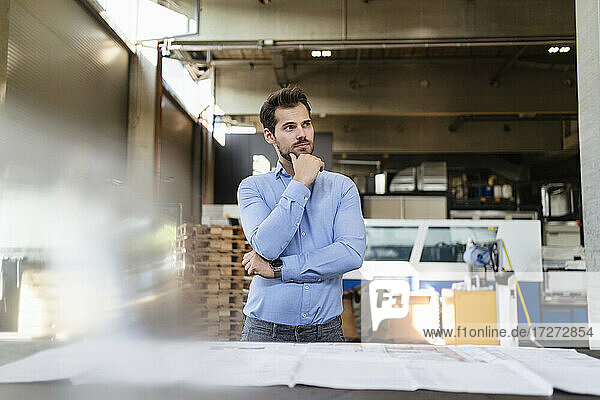 Thoughtful businessman with hand on chin standing by table at factory