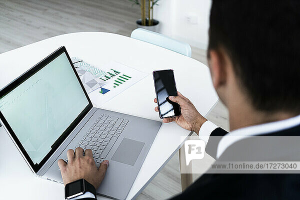 Geschäftsmann  der ein Mobiltelefon benutzt  während er mit einem Laptop am Schreibtisch in einem kreativen Büro sitzt
