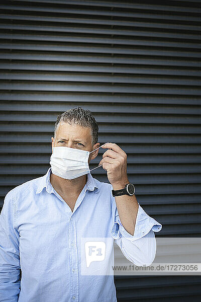 Mature man wearing protective face mask while standing against metal wall